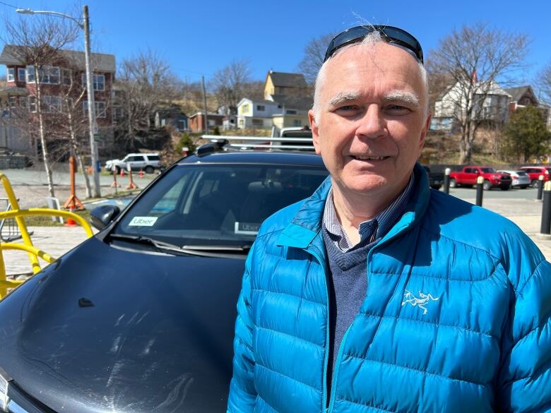 A man in a blue jacket standing in front of a black vehicle on a sunny day. 