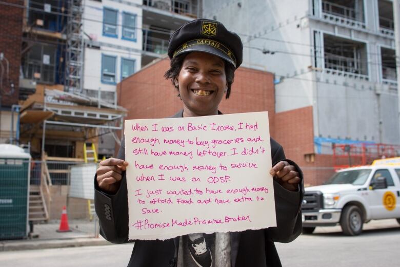 A person in a leather jacket and cap holds up a sign with text sharing how basic income improved their life. 