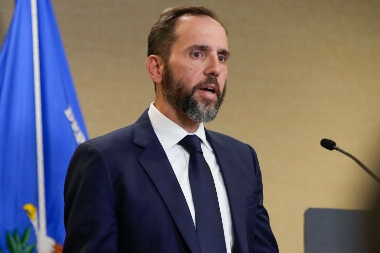 A bearded man in a suit and tie is shown speaking while standing in front of a flag.