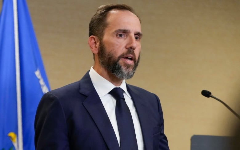 A bearded man in a suit and tie is shown speaking while standing in front of a flag.