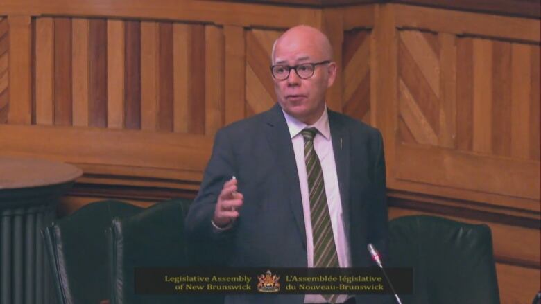 A man is standing and speaking in a room with wooden walls.