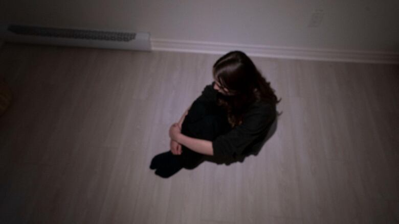 A girl sits on the floor with her hands grasped around her knees, her face not visible. 