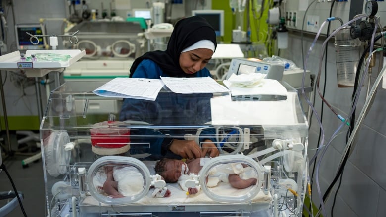 A woman wearing a headscarf in a hospital stands over an incubator containing an infant.