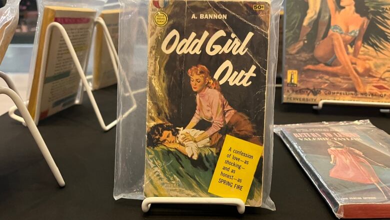 A pulp fiction novel, titled Odd Girl Out by Ann Bannon sits on a black table with other books in the background.