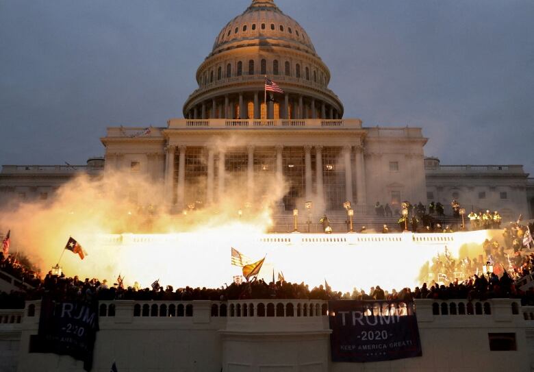 An august building is shown lit up, with many people gathered outside and flags waving in what appears to be a disruption.