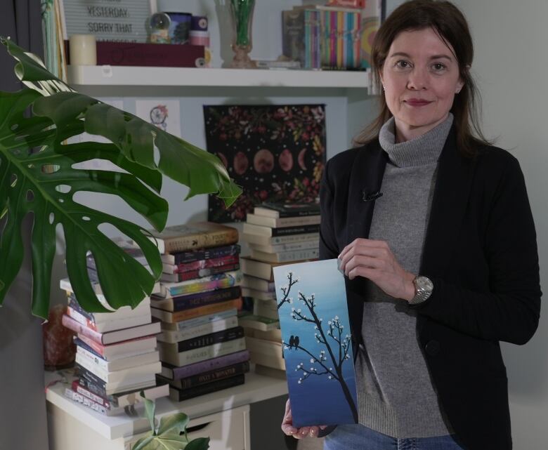 A woman indoors holds a small painting of a budding tree branch, with stacks of books and some plants seen behind her.