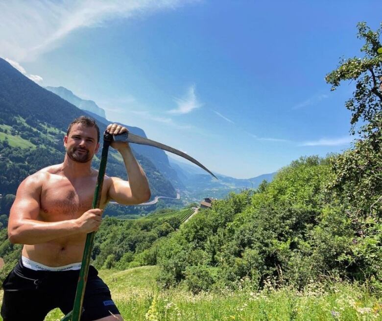 A muscular man stands shirtless on a mountainside while holding a scythe. 