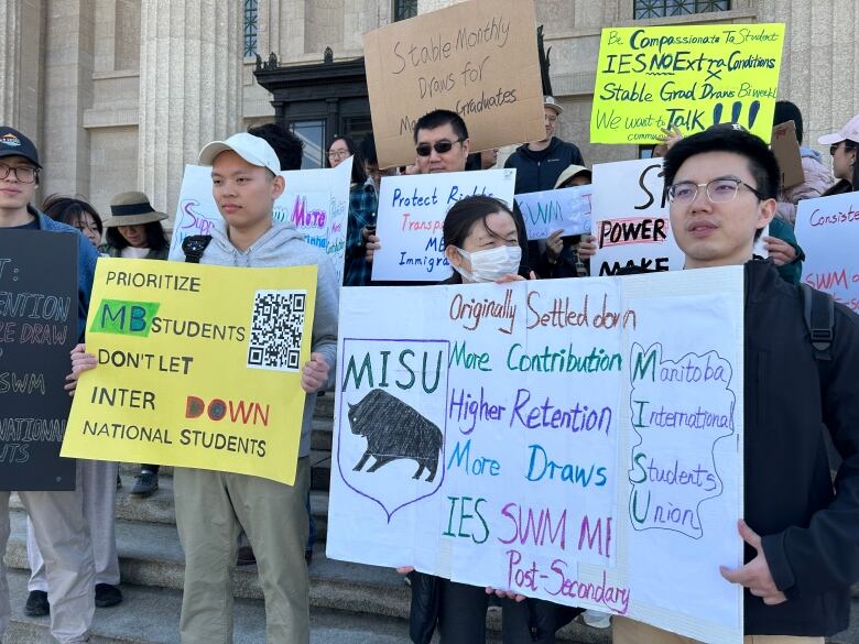 People demonstrating hold up signs with written messages, while standing on the steps of the legislature.