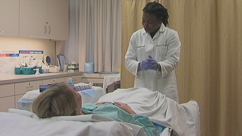 A physician in a white coat stands next to a woman lying on an examination table in a doctors office.