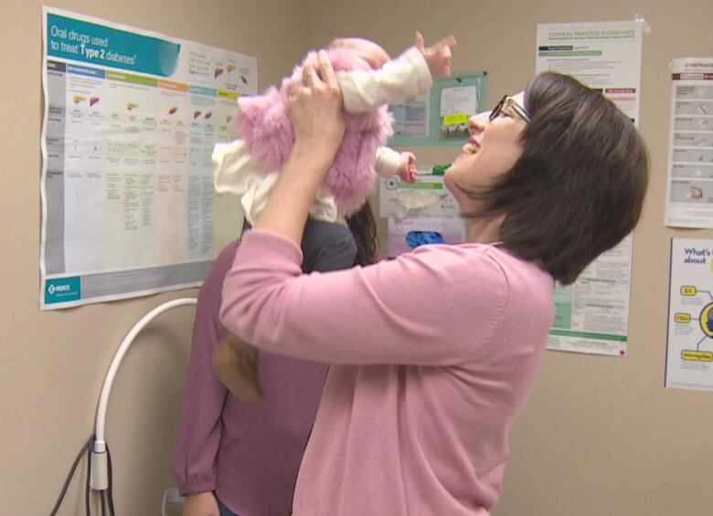 A doctor in a medical clinic smiles as she holds up a infant in a pink dress.