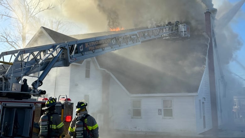 Two firefighters look on as smoke billows from the top of a large white building.