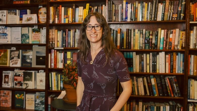 A woman in a plum frock stands in front of full bookshelves