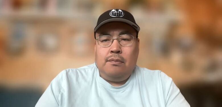 A man wearing a white t-shirt, glasses and a black Edmonton Oilers cap sitting down