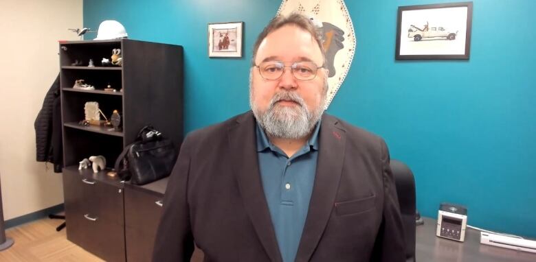 A man with a white beard and brown hair wearing a blue polo t-shirt under a black suit jacket and glasses standing in front of Inuit wall decorations