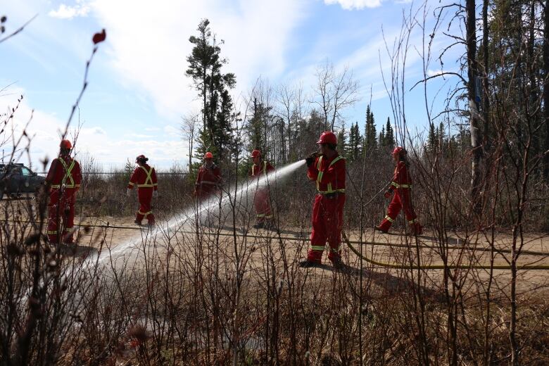 A group of men in red jumpsuits walk by the bush near a lake. They're all wearing red jumpsuits and hard hats. One man uses a firehose to spray water into the bush.