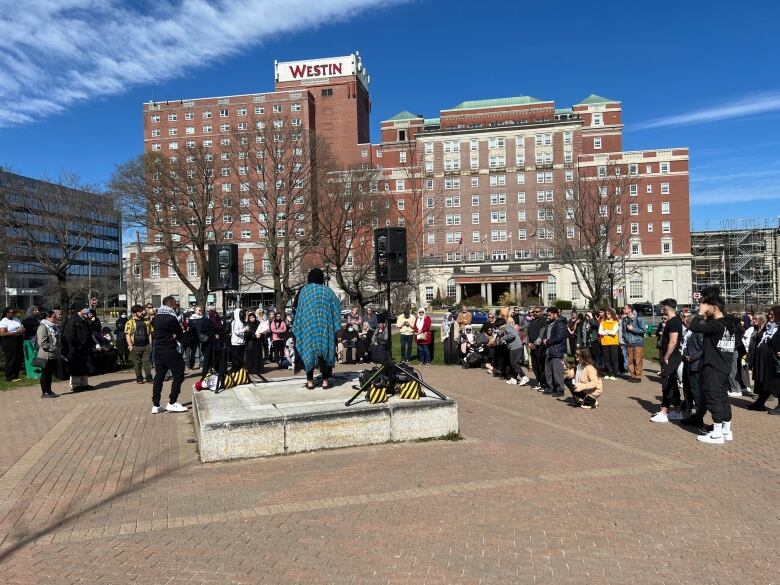 A large group of people stand around a speaker on a stage. The Westin hotel is in the background.