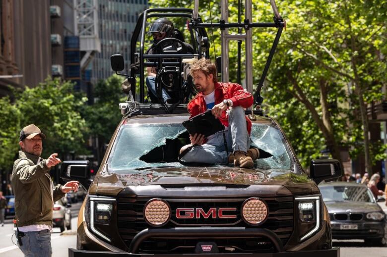 Ryan Gosling consults a tablet, while sitting on the hood of a truck with a shattered windshield.  To his left, director David Leitch is pointing off screen. 