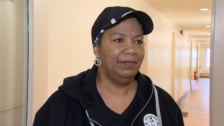 A woman stands in a hallway. She wears a black ballcap and black sweatshirt. She wears dangly, sparkly earrings. 