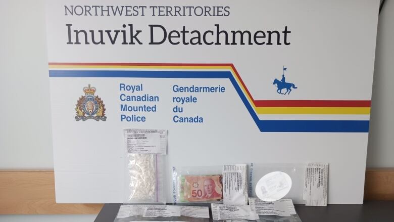 A small bag of cocaine, a small bag of cash and a few other items sit in front of an RCMP sign. 