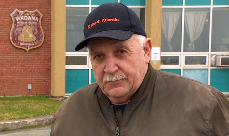 An older man wearing a brown jacket and black hat stands in front of a town hall. 