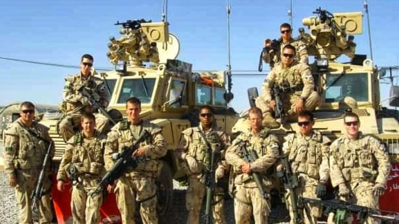 A group of soldiers wearing army fatigues is photographed in front of two armoured vehicles. 