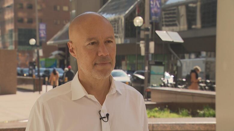 A bald man in a long sleeve white dress shirt stands in a small downtown park area with a busy street and glass buidling behind him in the distance.   