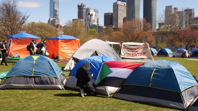 A person leans next to a tent with a Palestinian flag. Other people and tents are seen in the background. 