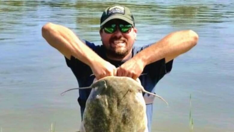 A man holds a monster catfish by the jaw with both hands on a riverbank. The head appears as wide as his torso. 