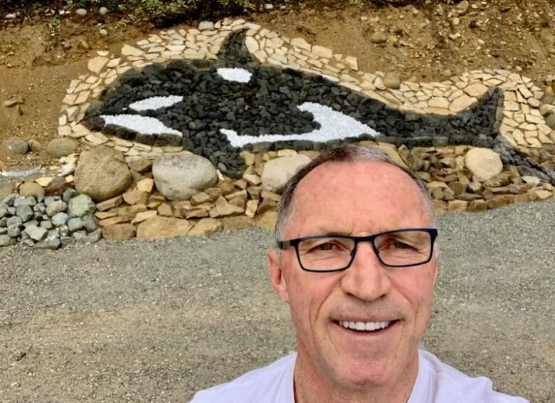 A close-up shot of a man with glasses. There's artwork featuring a black and white orca in the background crafted with rocks.