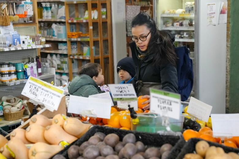 A woman with black hair wearing glasses and a dark coat stands behind a counter of produce.