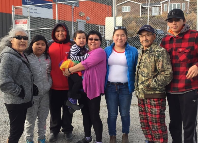 A group of people stand outside, side by side, in front of a building a chain-link fence. 