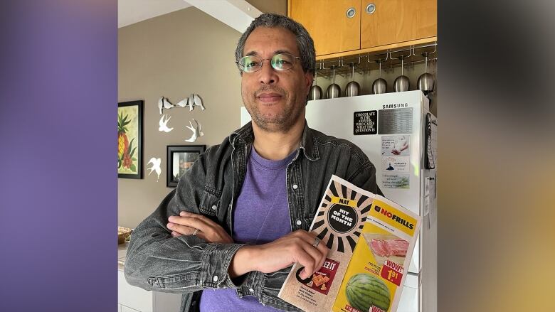 A man in a purple shirt stands with his arms crossed in a kitchen, holding a flyer that says No Frills.