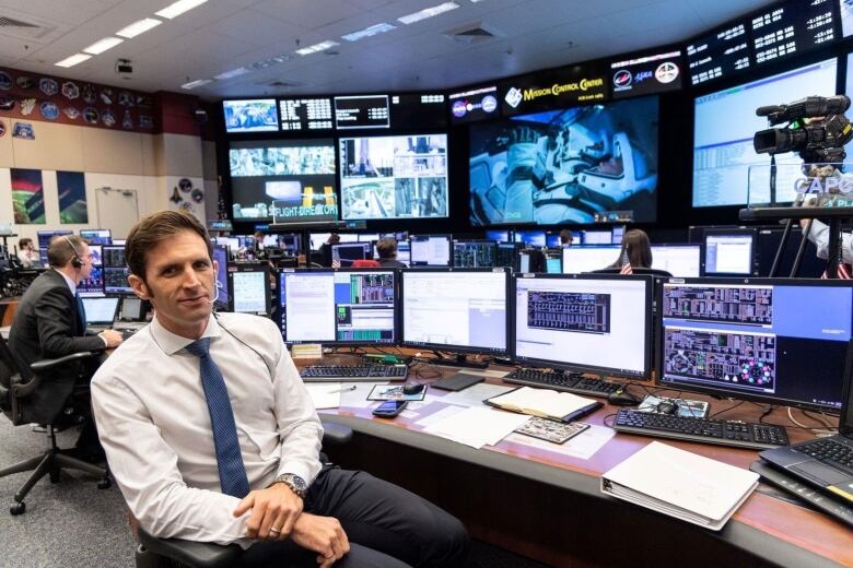 Man sits at busy work station in a white shirt and tie with numerous video screens in the background.