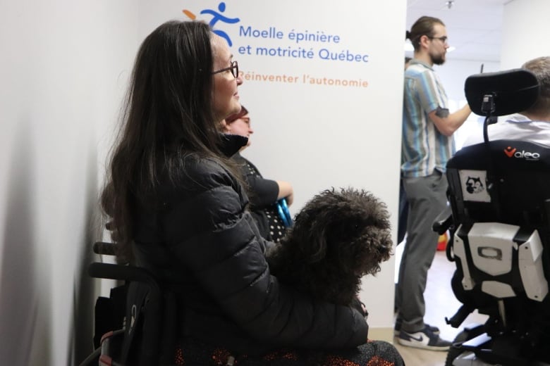 A woman sitting in a wheelchair smiles. Her small black dog sits on her lap.