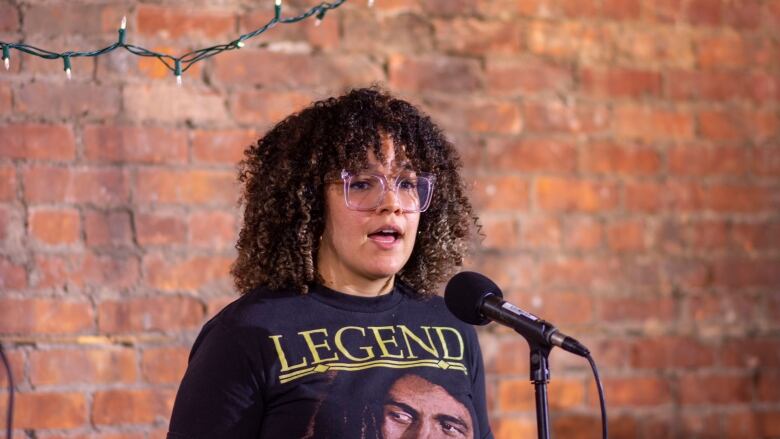 A person speaks into a microphone while standing in front of a brick wall.