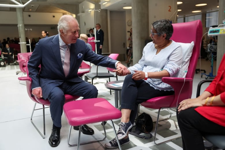 A seated person holds the hand of another person who is seated while receiving intravenous treatment.