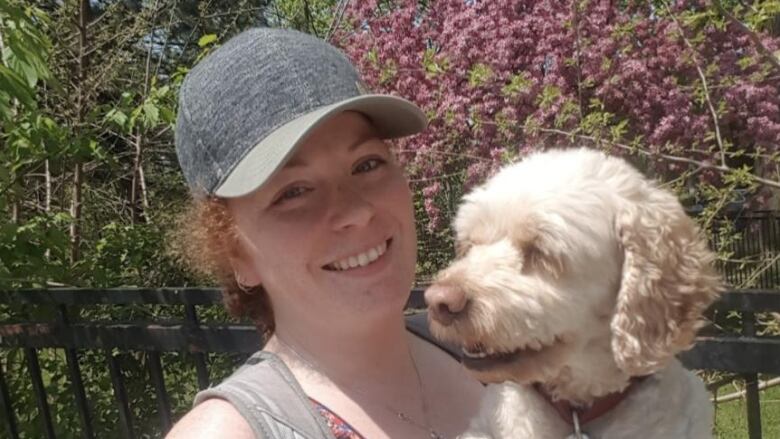 A red-haired woman in a blue, red and white tank top with anchors on it and a grey hat takes a selfie while holding a white fluffy dog.