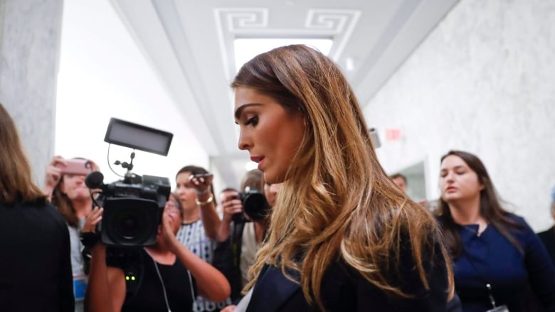 A woman in a black suit looks down as she walks past a row of cameras and reporters.