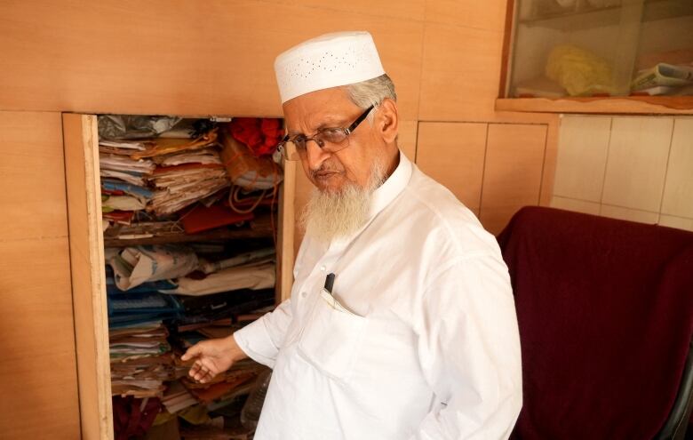 A man with a beard and white clothing shows a cabinet with stacks and folders of documents.