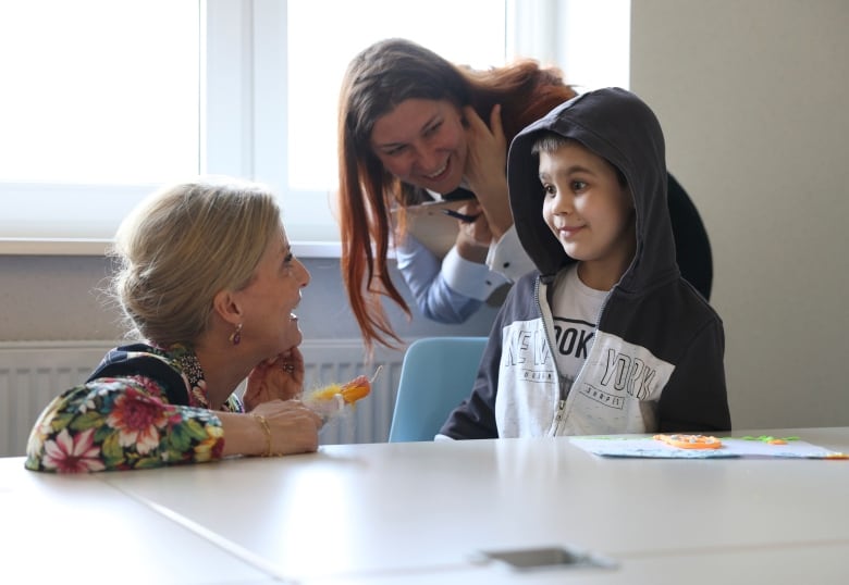 An adult kneels at a table while talking with a child and another adult.