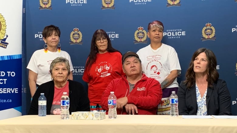 Two women and a man sit at a table as three women stand behind them in front of a backdrop written Edmonton police. 