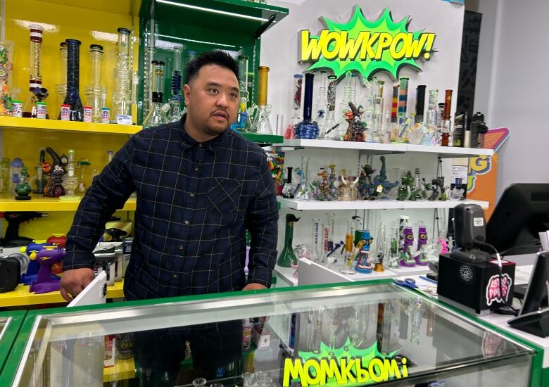 A man in a buttoned-up shirt is behind the counter of a cannabis store. A reflection of a green and yellow logo is shown on the glass table in front of him.