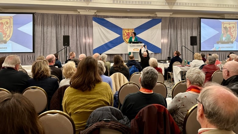 A crowd of seated people in foreground, look away from the camera toward a woman speaking at a podium, seen in the background.