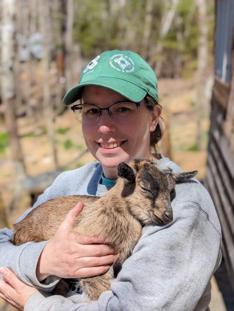 A smiling woman in a baseball cap cradles a baby goat in her arms.