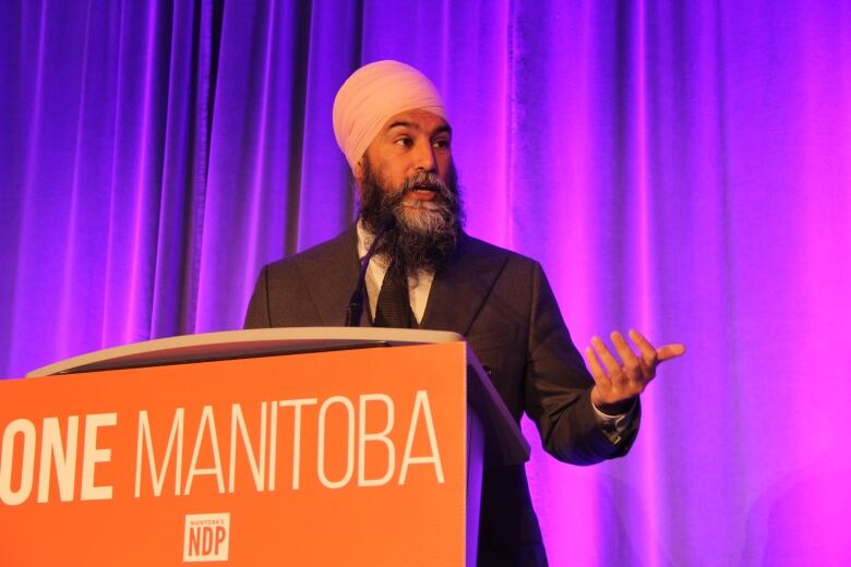 A man in a black suit and pink turban gestures with his hand to his left while standing in front of a blue curtain background.