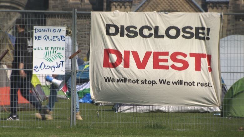 A sign hangs on a chain link fence that reads: Disclose. Divest. We will not stop. We will not rest.