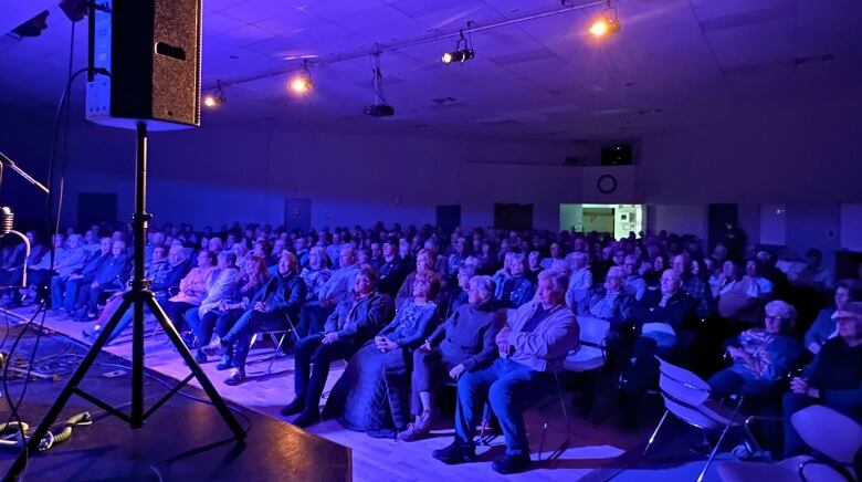 People fill a hall and appear purple under the stage lights.