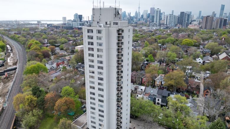 A tall apartment building is seen from a bird's eye view surrounded by trees, houses, a road and the Toronto skyline. 