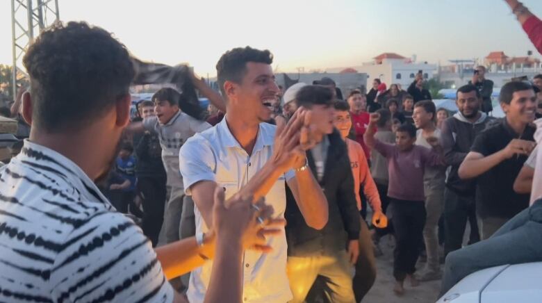 A man in a white T-shirt claps and smiles in a crowd of people celebrating good news at sunset.