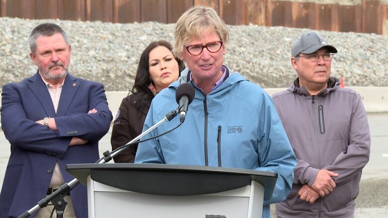 A woman stands at a podium outdoors speaking, with 3 people looking on from behind her.